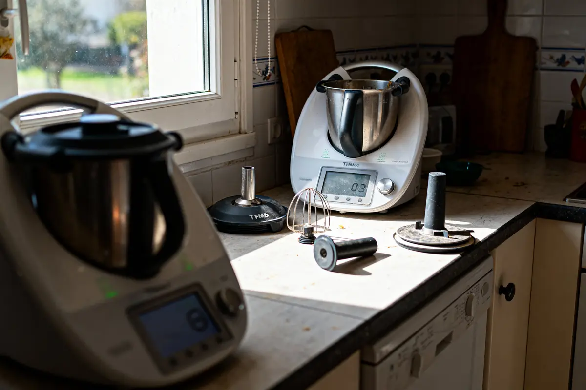 Deux robots de cuisine placés sur un plan de travail avec accessoires, près d'une fenêtre ensoleillée.