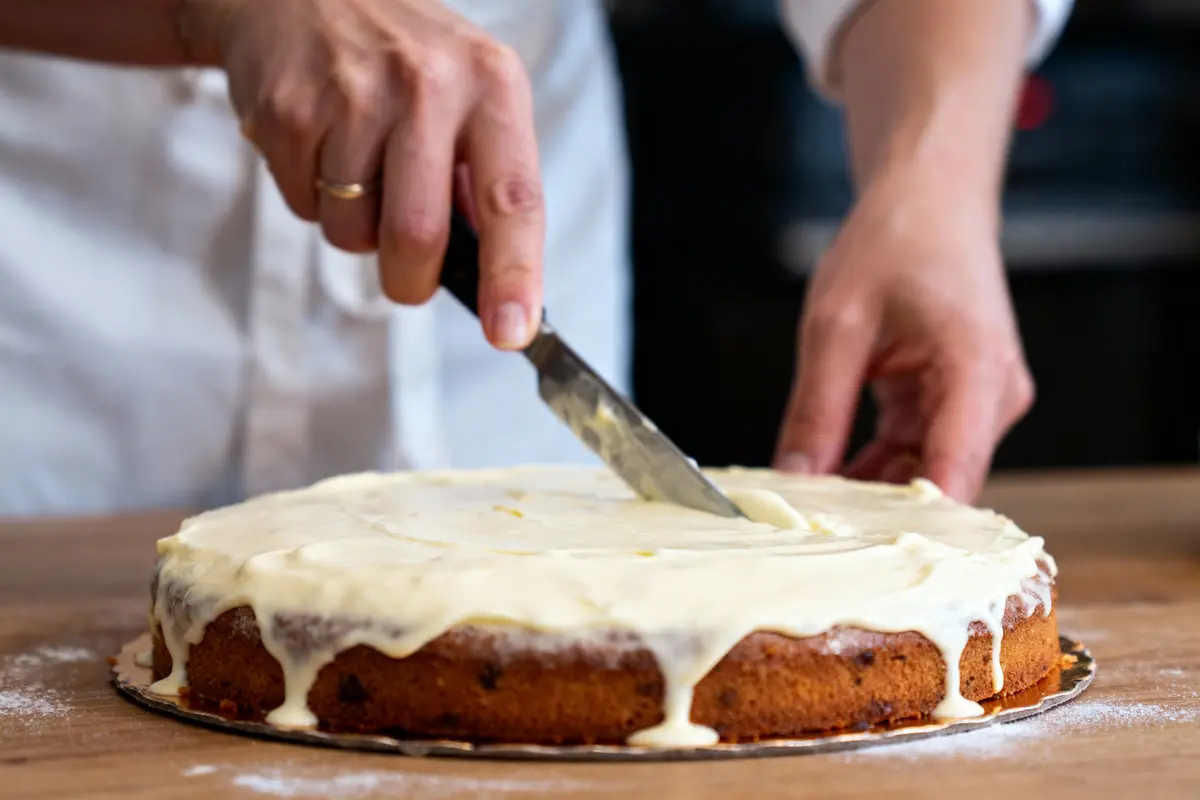 Personne étalant du glaçage blanc sur un gâteau rond à l'aide d'un couteau, sur une table en bois.