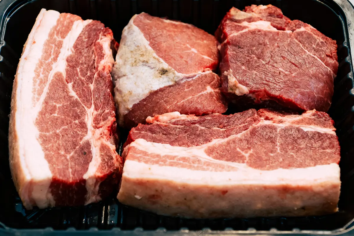 Quatre morceaux de viande crue rouge avec gras visibles dans un récipient en plastique noir.