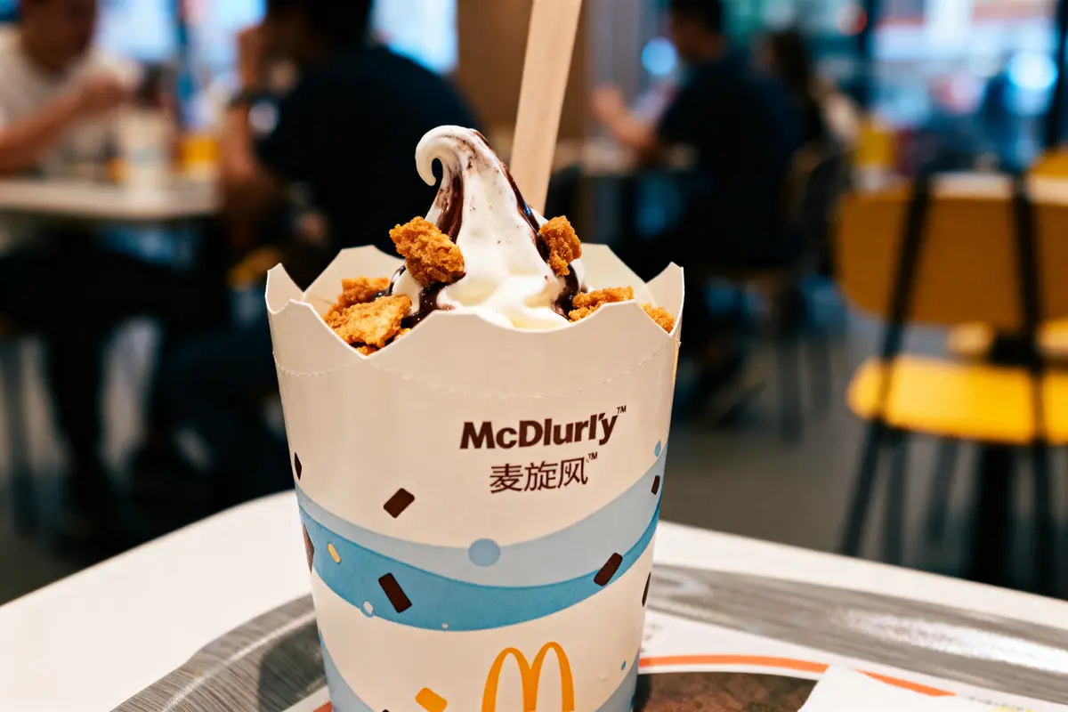 Crème glacée McDonald’s McFlurry avec garniture croquante, en intérieur flou de restaurant avec clients assis.