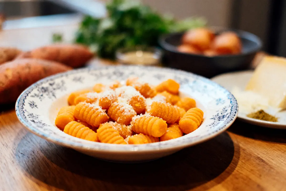 Assiette de gnocchis orange, saupoudrés de parmesan, sur une table en bois, avec ingrédients flous en arrière-plan.