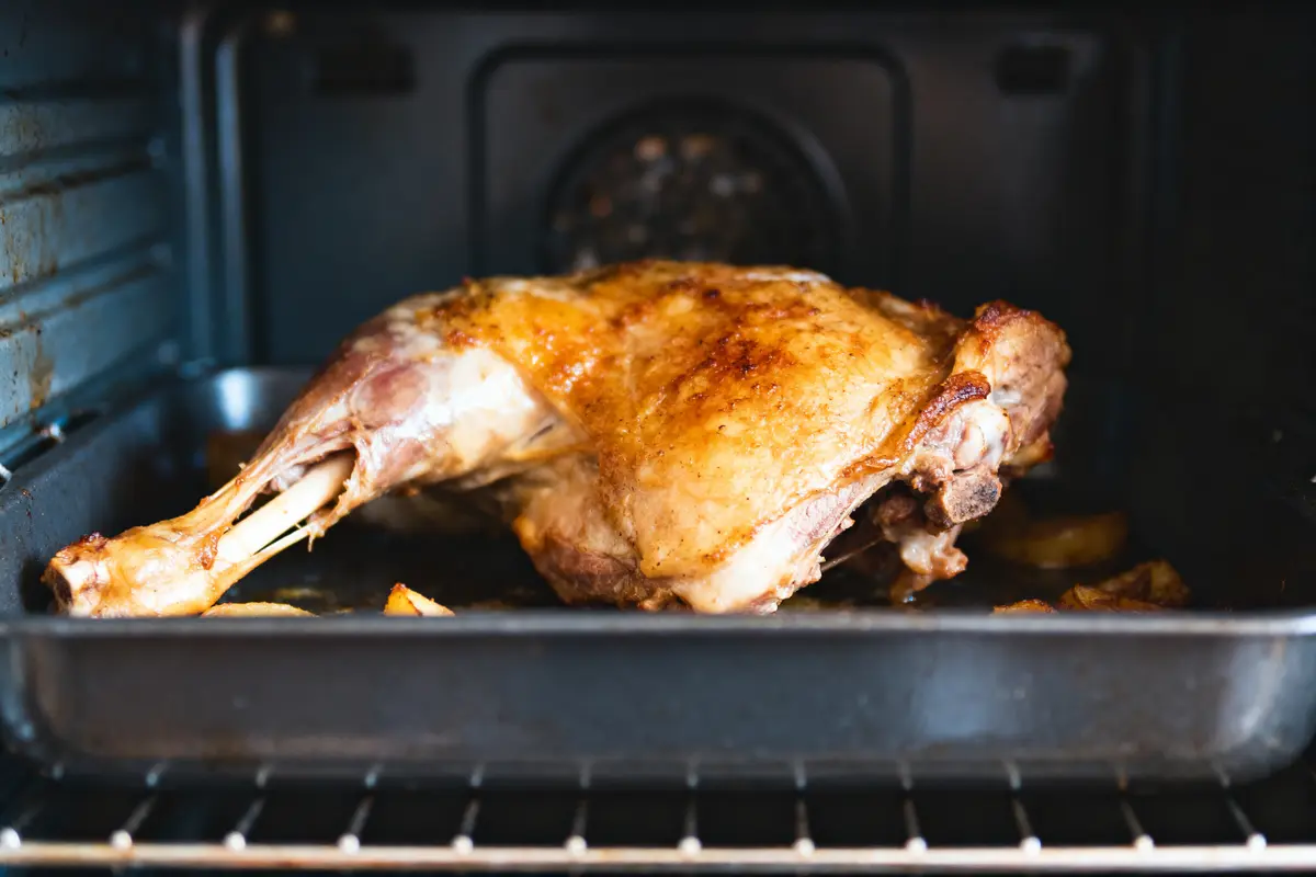Gigot d'agneau rôti dans un four, posé sur un plat noir avec pommes de terre, cuisson dorée.
