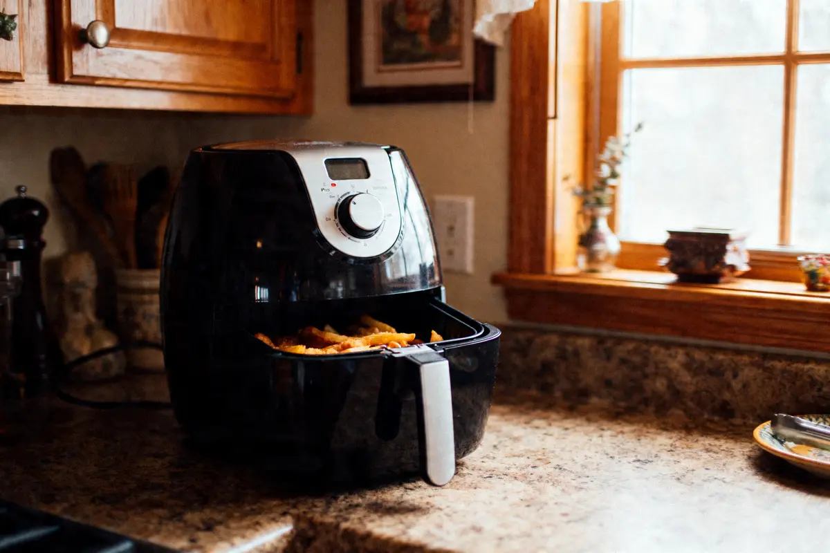 Friteuse à air sur plan de travail de cuisine avec frites, lumière naturelle par fenêtre derrière.