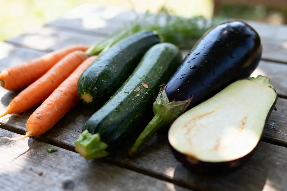 Carottes, courgettes et aubergines sur une table en bois, éclairage naturel, légumes frais et entiers.