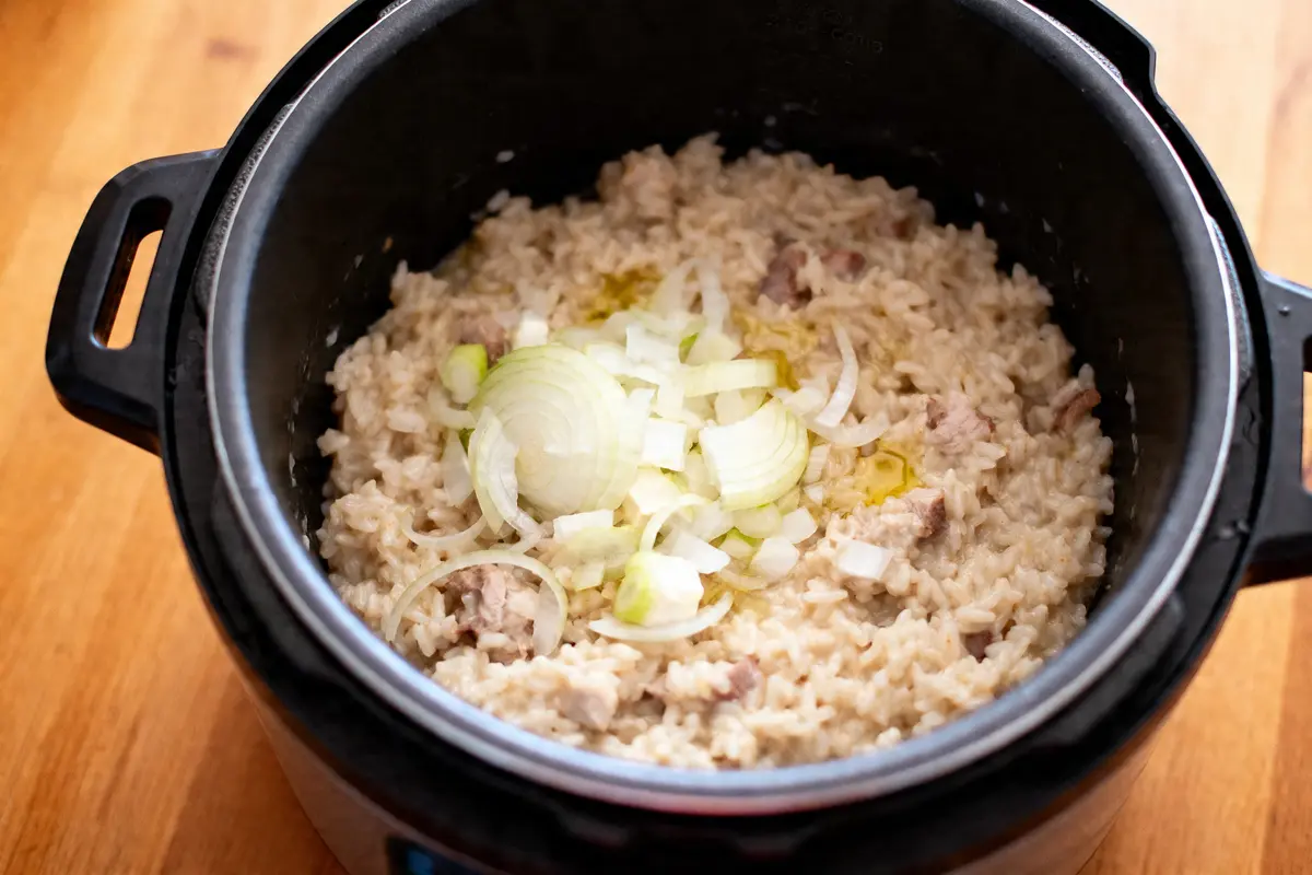 Riz crémeux avec oignons émincés, huile et morceaux de viande dans un autocuiseur noir.
